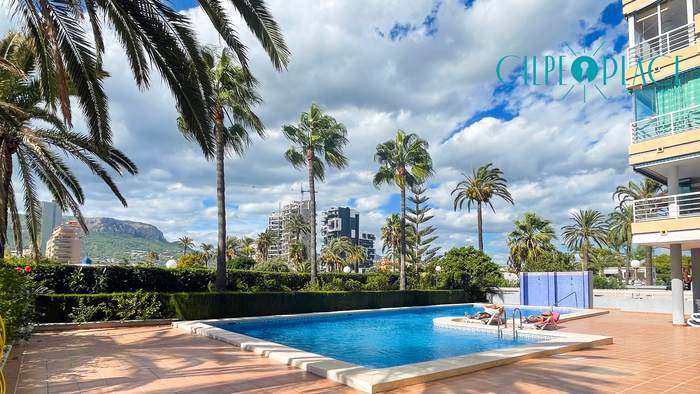 Piscina comunitaria de Oasis rodeada de palmeras, con el Peñón de Ifach al fondo